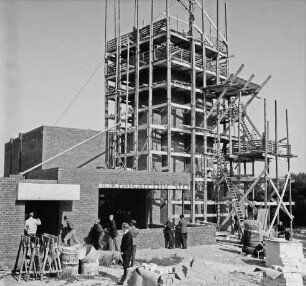 Evangelische Kirche: Rohbau: Kirchturm mit Gerüsten: Richtfest: davor Gemeindemitglieder: im Vordergrund Baumaterial