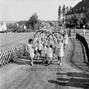 Kindervogelschießen: Umzug: durch Grünanlage zwischen Matthias-Claudius-Schule (hinten rechts) und Ahrensböker Straße (hinten links): Mädchen mit Blumenbögen, Jungen mit Blumenstäben: beiderseits Jägerzaun, 29. Juni 1963