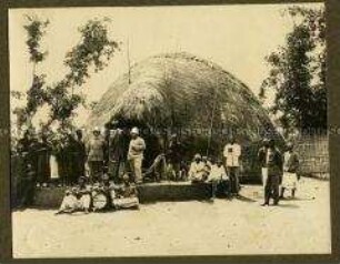 Europäer mit einem afrikanischen Häuptling und Gefolge vor dessen Hütte