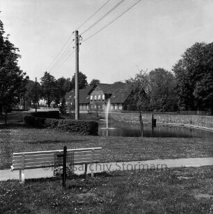 Dorfstraße: Dorfteich mit Springbrunnen: vorn Grünanlage mit Weg, Bank und Papierkorb: links Strommasten: hinten Häuser und Bäume