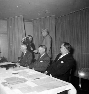 Keck, Erika: Kreispräsidentin: Kreisberufschule Bad Oldesloe: Kreistagssitzung am 18. Mai 1955: Verabschiedung und Überreichung eines Blumenstraußes durch Landrat Wilhelm Siegel: Präsidiumstisch mit dem neuen Kreispräsidenten Friedrich Hardt und den Stellvertretern Willy Esser und Paul Manthey: hinten Vorhänge