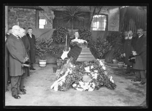 Aufbahrung eines Toten im geschlossenen Sarg mit Blumenschmuck und Kranz, Pfarrer hinter dem Sarg, Personen links und rechts