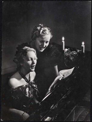 o. T. (Klavierkonzert) : Zwei Frauen beim Musizieren, die eine am Flügel sitzend und singend, die andere hinter ihr stehend und singend, auf dem Flügel ein Gesteck mit zwei brennenden Kerzen.