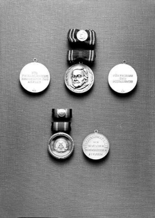 Zwei Medaillen der Deutschen Demokratischen Republik (DDR). Oben: Clara-Zetkin-Medaille an Spange (Silber; 36 mm Durchmesser. 1954 gestiftet) inklusive Interimsspange. Avers (Mitte) mit Portrait Clara Zetkins und Revers mit Inschrift (links erste Form 1954/1958, rechts zweite Form 1959/1972). Unten: Verdienstmedaille der DDR (versilbert, emailliert; 33 mm Durchmesser. 1959 gestiftet) an Spange (11 x 24 mm) inklusive Interimsspange. Avers (rechts) und Revers mit Inschrift