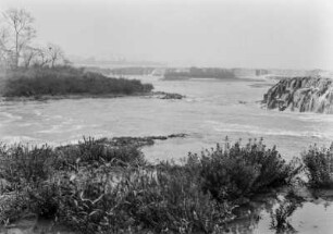Flusslandschaft des Rio Grande : Eine Flusslandschaft des Rio Grande mit Felsen im Flusslauf, Gras- und Buschvegetation am Ufer und Wasserfällen im Hintergrund.