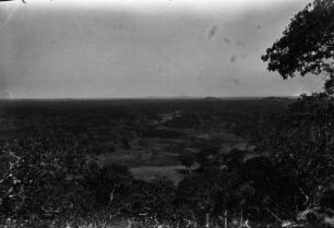 Hügellandschaft : Quazithügel auf einer Ebene. Am Horizont zwei Gneisdome. Im Bildvordergrund ein Dambo im Wald mit abgebrannten Grasflächen.