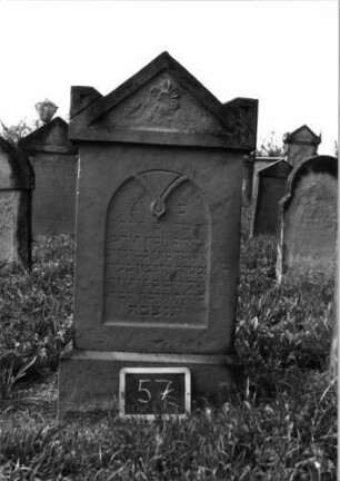 Grabstein 57 (Index: 99937000) Material: Sandstein Zustand: verwittert Maße HxBxT: 130x58x14 cm Beschreibung: Stele, Sockel, Giebel mit stilisierten Eckakroteren, Schriftfeld eingetieft mit Rundbogenabschluß Symbolik: Blüte im Giebel, stilisiertes Maßwerk mit herabhängender Blüte: Grabstein 57
