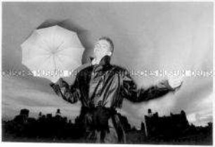 Junger Mann in schwarzem Mantel und ausgebreiteten Armen hält einen aufgespannten Regenschirm in die Höhe (Sonderthema: Traum-Bilder)