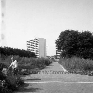 Siedlung Ehmkenberg II: Fußwege mit seitlichem Gebüsch, links ein Fußgänger, im Hintergrund die Wohnhochhäuser der Straße Im Hölk