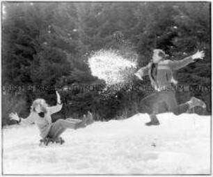 Springender Mann wirft Schnee auf eine Frau, die auf einem Schlitten sitzt (Altersgruppe bis 14)
