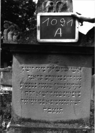 Grabstein 1091 (Index: 100104145) Material: Sandstein Zustand: teilweise angewittert Maße HxBxT: 178x53x20 cm Beschreibung: Stele auf getrepptem Sockel, Aufsatz mit Dreiecksabschluss und dahintergestellten spitzbogigen Giebel- sowie Eckakroterien, Giebelakroter mit Mohnkapseln, Eckakroterien mit Voluten, unterster Sockel felsartig gearbeitet, Stele wirkt unproportioniert;Mittelteil ist zu niedrig im Verhältnis zum Sockel Symbolik: Mohnkapseln, Voluten: Grabstein 1091A