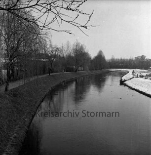 Trave: rechts Travewanderweg mit Personen, Bäumen, Zaun, Häuser und Schuppen: hinten rechts Wiese: dahinter Gewerbegebäude: Winter