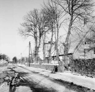 Dorfstraße mit Kopfsteinpflaster: rechts Bauerngehöft mit Silo: davor Vorgarten und Baumreihe: vorn Kind mit Schlitten: Winter