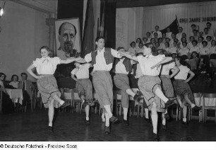 Tänzer in Tracht der Leipziger Eisen- und Stahlwerke, im Hintergrund ein Chor sowie ein Porträt Walter Ulbrichts
