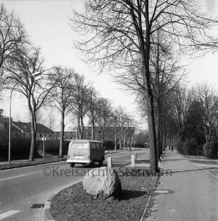 Sieker Landstraße: rechts Fußgängerweg: vorn Grünfläche mit Findling: dahinter Baumreihe: links VW-Bus: dahinter weitere Baumreihe und Gebäude