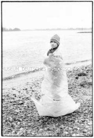 Kleiner Junge mit Mütze steht mit einer Plastikplane verhüllt an einem Flussufer (Altersgruppe 18-21)