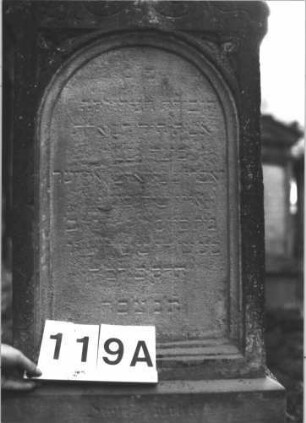 Grabstein 119 (Index: 99898918) Material: Sandstein Zustand: stark verwittert Maße HxBxT: 150x53x24 cm Beschreibung: aufgesockelte Stele mit Konvexbogenabschluss, eingeritzte Mittelpalmette, Voluten und Ranken im Bogenfeld, Unterer Sockel felsartig gearbeitet Symbolik: Palmette, Ranken, Voluten: Grabstein 119A