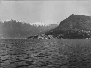 Am See : Blick über eine See und das andere Ufer mit einer Stadt und hohen Bergen im Hintergrund