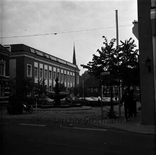Markt: vorne die Hagenstraße, mittig der Gänselieselbrunnen, dahinter parkende Fahrzeuge, links das Stadthaus, vorne rechts Passant auf dem Gehweg, im Hintergrund Gebäude der Hindenburgstraße, Blick Richtung Süden
