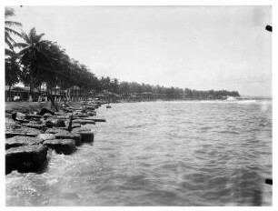 Colón, Panama. Blick in die Bucht entlang des Palmenstrandes mit großen Steinblöcken als Wellenbrecher, im Hintergrund Wohnhäuser oder Strandhäuser