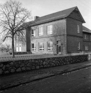 Volksschule mit Erweiterungsbau: links Schulhof mit Baum: davor Mauer mit Zaun: im Vordergrund Dorfstraße