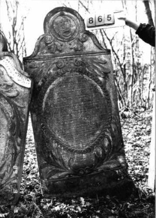 Grabstein 865 (Index: 99751227) Material: Sandstein Zustand: leicht verwittert Maße HxBxT: 171x77x9 cm Beschreibung: Stele, geschwungen eingezogener Rundbogenabschluß, ovales Schriftfeld Symbolik: Levitenkanne mit Schale, Ranken, Blätter, Scheibenfries, Voluten: Grabstein 865