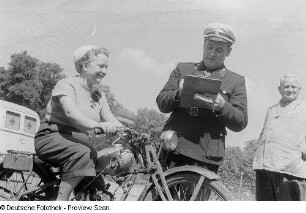 Eine Frau auf einem Motorrad, ein Polizist mit einem Klemmbrett sowie ein weiterer Mann, im Hintergrund ein Krankenwagen