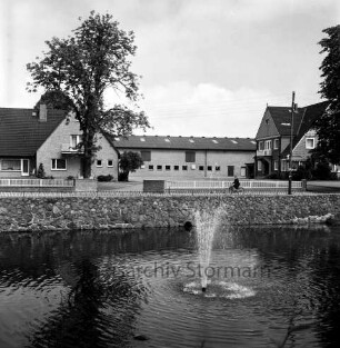 Dorfstraße: vorn Dorfteich mit Springbrunnen: hinten Häuser und Bäume