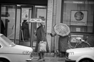 Paul-von-Schoenaich-Straße: zwei Frauen mit Regenschirmen vorn Geschäftsstelle der Kreissparkasse Stormarn: davor Pkws und Fahrrad