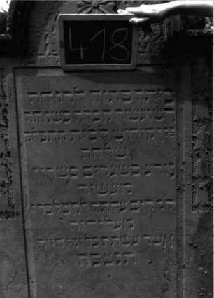 Grabstein 418 (Index: 100102786) Material: Sandstein Zustand: stark verwittert, abgeblättert, teilweiser Textverlust Maße HxBxT: 169x82x13 cm Beschreibung: Stele, eingezogener Rundbogenabschluss mit Verdachung, im Bogen Blüte, auf den Einzügen Kugelaufsätze, Schriftfeld rechteckig eingetieft, auf dem Rahmen Blütenfries Symbolik: Blütenfries: Grabstein 418 (Detailaufnahme 2)