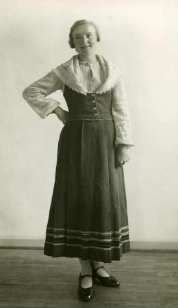 Frau in Volkstracht mit handgewebtem Kleid. Stehend, mit in der Taille gestützem rechten Arm. Frontal. Halbtotale