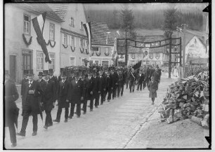 Primizfeier Heinzelmann in Stetten unter Holstein; Prozession durch einen geschmückten Torbogen; im Mittelpunkt Männer mit Zylinder mit Orden geschmückt