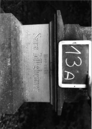 Grabstein 13 (Index: 100090868) Material: Sandstein Zustand: Schrifttafel/Aufsatz fehlt Maße HxBxT: 160x43x28 cm Beschreibung: Stele, getreppt. Sockel, eingezog. Rundbogenabschl. m. Veradchung, Schriftfeld rechteck Symbolik: nach unten hängender Blumenstaruß: Grabstein 13A