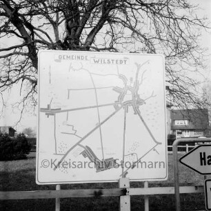 Dorfring: Schild mit Ortsplan: rechts Verkehrsschild: dahinter Friedenseiche und Grünanlage am Ehrenmal 1939-1945: hinten rechts und links Gebäude