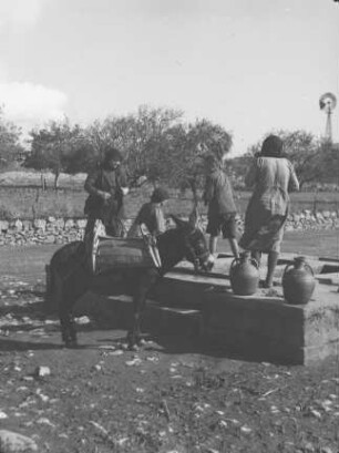 Frauen am Brunnen : Drei Frauen und ein Junge an einem Dorfbrunnen; im Hintergrund ein Windrad.