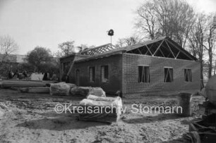 Dorfgemeinschaftshaus: Dorfstraße: Richtfest: Rohbau mit Richtkrone: hinten links Gäste: vorn Bretterstapel, Paletten mit Baumaterial, Tonne und Baumaschine: hinten Bäume