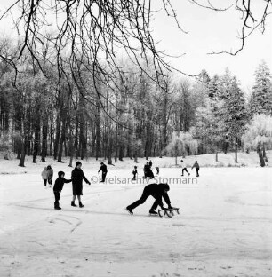 Kurpark: Salzteich: vorn Junge mit Schlittschuhen und Schlitten: links Frau mit Kind auf Eisfläche: hinten mehrere Kinder mit Schlittschuhen und Schlitten auf Eisfläche: dahinter Bäume: Winter