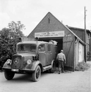 Freiwillige Feuerwehr: Hauptstraße: Feuerwehrgerätehaus mit Löschfahrzeug und Mann: dahinter Bäume, Haus und Strommast