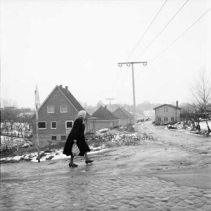 Neubausiedlung am Burdieksbach: vorn links Frau mit Einkaufsbeutel: hinten rechts Strommast: daneben altes Haus: Winter