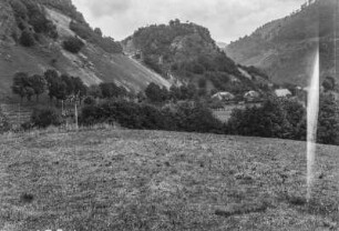 Dorf : Blick auf ein Dorf in einer Hochebene. Im Hintergrund bewaldete Berghänge, im Vordergrund Wiese
