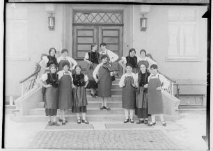 Haushaltungsschule in Gammertingen 1931; Junge Frauen mit Schürzen bei einer Aufführung mit Geigenspielerin auf einer Treppe vor dem Hauseingang