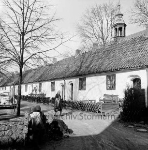 Gottesbuden: südliche Reihe: Jägerzaun, Ruhebank, Handwerker an Tür: davor alte Frau auf Weg, Auto, Wäscheleine: rechts dahinter Dachreiter von Schlosskirche: im Vordergrund zwei Kinder