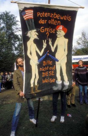 Ein Demonstrant hält ein Plakat, auf dem ein alter amerikanischer und ein alter russischer Mann ihre Waffen zeigen: Potenzbeweis der alten Greise? Bitte nicht auf diese Weise