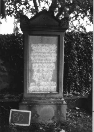 Grabstein 80 (Index: 100080577) Material: Sandstein, weiße Marmorplatte Zustand: verwittert Maße HxBxT: 141x50x28 cm Beschreibung: profiliert. Sockel, Giebel m. stilisiert. Eckakroteren, rechteck. Schriftfeld eingetie Symbolik: Sternchen im Giebel, Blätter in den Akroteren: Grabstein 80
