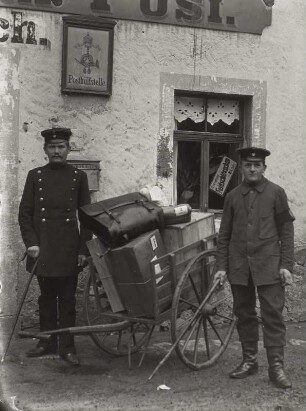 Postbeamter und Gehilfe mit Paketkarre vor einer ländlichen Poststelle