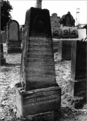 Grabstein 294 (Index: 99797834) Material: Sandstein, weiß Zustand: stark verwittert Maße HxBxT: 109x35x20 cm Beschreibung: Obelisk, oben abgebrochen, Sockel Symbolik: Blütenkranz: Grabstein 294