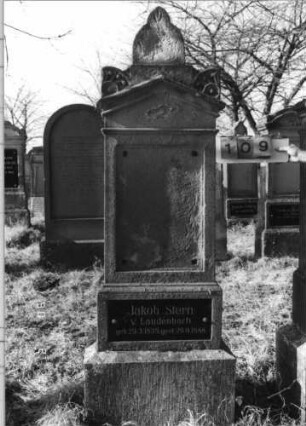 Grabstein 109 (Index: 100116424) Material: Sandstein, Granitplatte Zustand: Schriftplatte an der Stele fehlt, sonst leicht verwittert Maße HxBxT: 212x54x28 cm Beschreibung: doppelt aufgesockelte Stele mit Giebelabschluß, Eckakroteren und Aufsatz, eingesetzte Schriftplatten auf Stele und oberem Sockel: Grabstein 109