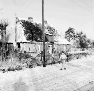 Reetdachkate an Möllner Landstraße mit Vorgarten, Hecke und Zaun: davor Kind und Strommast: Winter