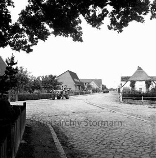 Schlamersdorf: Segeberger Straße: vorn Dorfstraße mit Kopfsteinpflaster: rechts Feuerlöschteich mit Geländer: links Einfriedung des Ehrenmals mit Baumästen: hinten links Trecker: hinten rechts landwirtschaftliches Fuhrwerk, Wohnhäuser und Bauernhäuser
