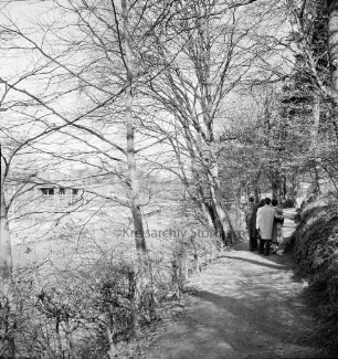 Trave: vorn Wanderweg mit Baumreihe am Ufer: links zwei Paare: hinten links am gegenüberliegenden Ufer Badeanstalt: hinten rechts Brücke